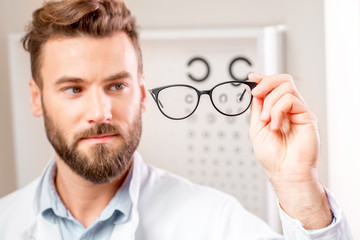 Handsome ophthalmologist looking at the glasses in front of the eye chart in the cabinet. Image with small deph of field focused on hands and glasses