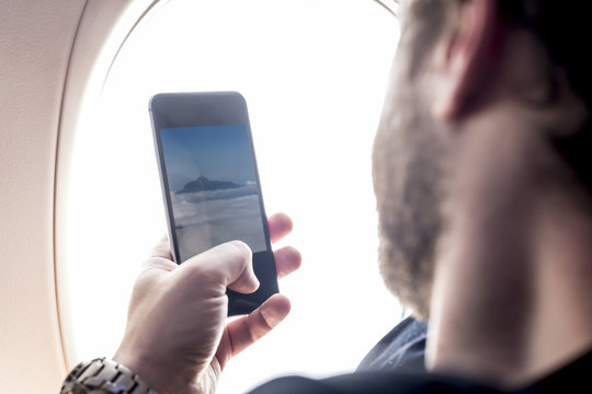 Bearded man sitting at airplane window and taking photo on telephone