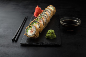 Delicious fried sushi roll with salmon, avocado, tofu cheese on a black board on a dark background.