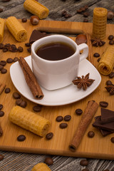 Small white cup of coffee, cinnamon sticks, cocoa beans, star anise, hazelnuts, chocolate, and cookies on wooden background