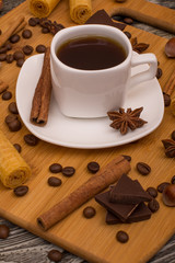 Small white cup of coffee, cinnamon sticks, cocoa beans, star anise, hazelnuts, chocolate, and cookies on wooden background