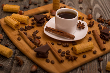 Small white cup of coffee, cinnamon sticks, cocoa beans, star anise, hazelnuts, chocolate, and cookies on wooden background