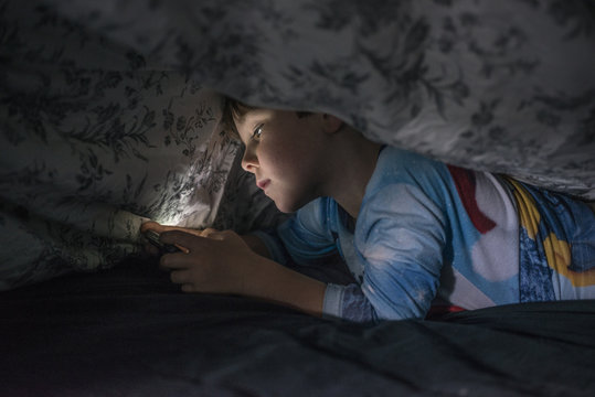 Young boy playing with cell phone under duvet