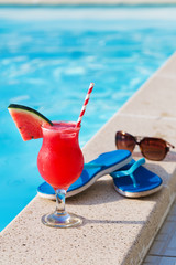 Glass of orange juice drink fresh with flowers, sunglasses and slippers on border of a swimming pool - holiday tropical concept