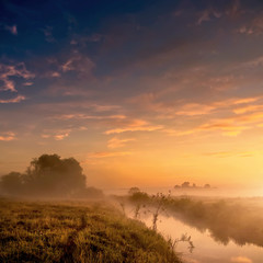 Fantastic foggy river with fresh grass in the sunlight. majestic misty sunrise with colorful clouds on the sky,    Dramatic unusual scene. Warm sundown over meadow. Beauty in the world.