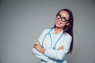 Smiling young woman doctor in glasses and white coat standing on grey background. Hands folded in front of her