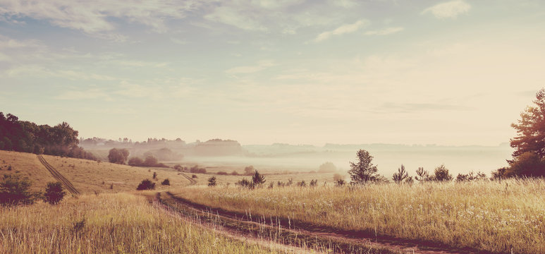 Fantastic foggy sunny day. ground road in the rural field with fresh grass in the sunlight. majestic misty sunrise with colorful clouds on the sky, Dramatic picturesque scene. vintage creative effect