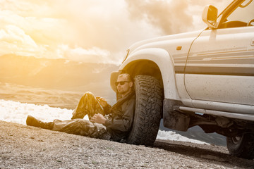 Man sits on top of a mountain next to the wheel  an SUV and  rest. Against the backdrop  the snow  the mountains. The height  3000 meters. Altai, Russia.