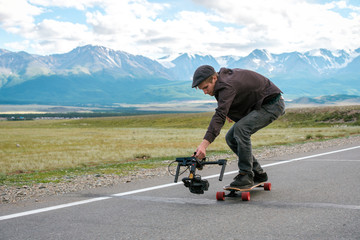 Man rides a longboard and shoot camera with electronic steadycam. Long road in the field on  background of mountains  glaciers.