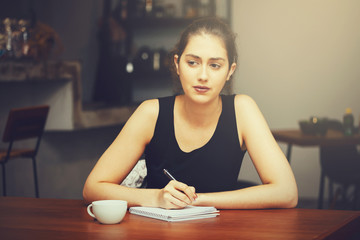 Young Caucasian woman thinking and writing something on a notebook in cafe