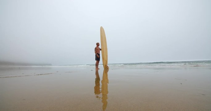 Mature Surfer Stands with Surfboard Looking out to the Ocean Waves.