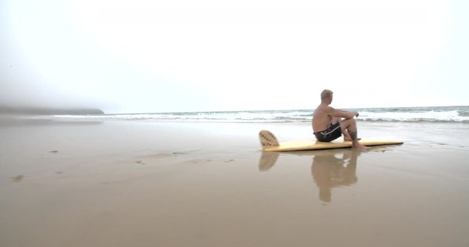 Mature Surfer sits on his Surfboard Looking out to the Ocean Waves.