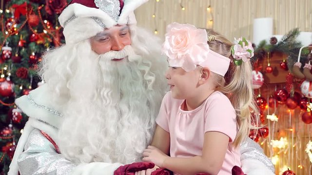 Beautiful little girl having fun smiling Santa Claus and sitting on his lap.
