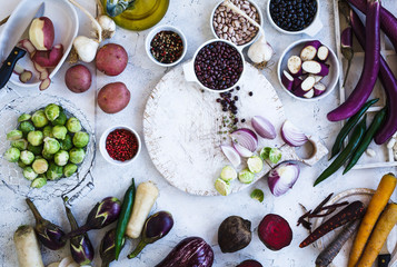 Winter food ingredients. Variety eggplants, colorful carrots, beetroot, potatoes, black and red beans, brussels sprout, herbs and spices on chopping board, over rustic white table. Top view. 
