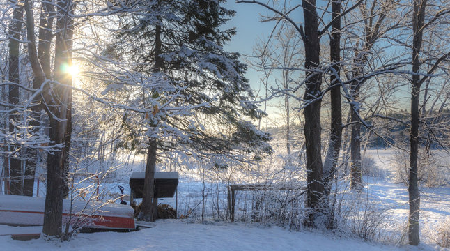 Fototapeta Winter sunrise morning in a cottage back woods.  Wintry fresh fallen snow is revealed as the sun comes up on cold, wooded cottage lake.  Upside down boat waits for the next season.