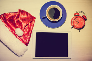 Santa Claus hat, tablet pc computer, coffee cup and alarm clock on white wooden table background