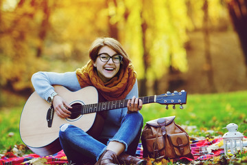 Fototapeta na wymiar Outdoor full body portrait of a young beautiful happy smiling girl playing guitar in the park. Model looking at camera. Lady wearing stylish knitted autumn clothes. Toned. Copy space for text