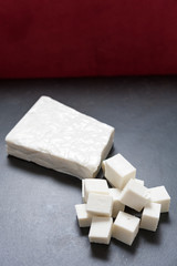 Buffalo paneer Indian cheese on black slate cutting chopping board counter in one large piece and smaller cubes with dark red background and copy space above