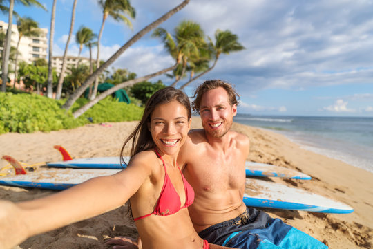 Happy young interracial couple taking phone selfie picture relaxing sitting near surfboards on beach holiday having fun doing surfing activity class. Travel vacation destination watersports.