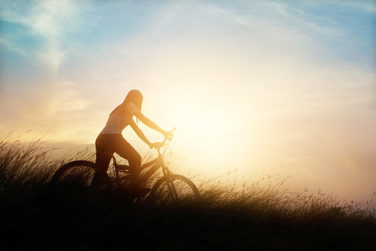 Woman with bicycle on a rural road with grass at sunset background