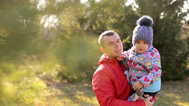 Dad in a red jacket holding his little daughter on his arms. Family walks in the woods. Sunny weather.