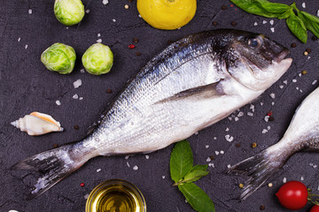 Raw fresh dorado fish with brussels sprouts, tomatoes, lemon, young potato, greens, bread, white wine bottle and olive oil on dark background. View from above, top studio shot