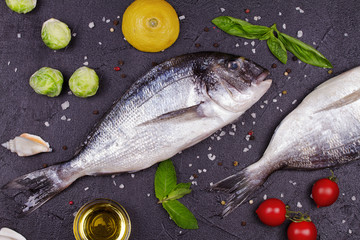 Raw fresh dorado fish with brussels sprouts, tomatoes, lemon, young potato, greens, bread, white wine bottle and olive oil on dark background. View from above, top studio shot
