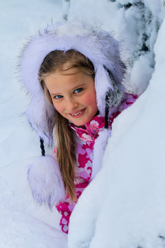 Portrait of child girl in winter clothes with falling snow near tree. Warm hat and lots of snow.