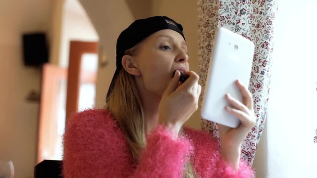 Girl in baseball cap using tablet as a mirror while painting her lips
