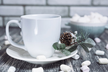 Hot chocolate with marshmallows. Drink in a white ceramic cup with zephyr on wooden table. Decorative pine branch with the cone on plate, saucer