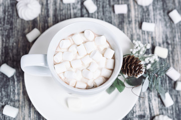 Fototapeta na wymiar Hot chocolate with marshmallows. Drink in a white ceramic cup with zephyr on wooden table. Decorative pine branch with the cone on plate, saucer.