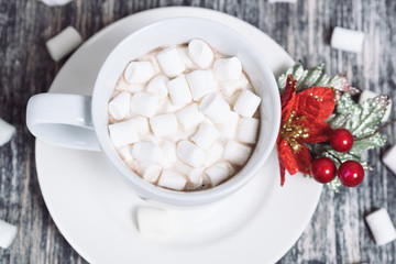 Obraz na płótnie Canvas Hot chocolate with marshmallows. Drink in a white ceramic cup with zephyr on wooden table. Red poinsettia flower on plate, saucer. 