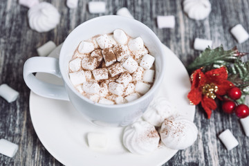 Hot chocolate with marshmallows. Drink in a white ceramic cup with zephyr on wooden table. Red poinsettia flower on plate, saucer. 