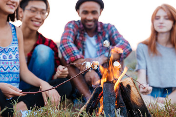Cheerful young people talking and preparing marshmallows on bonfire outdoors