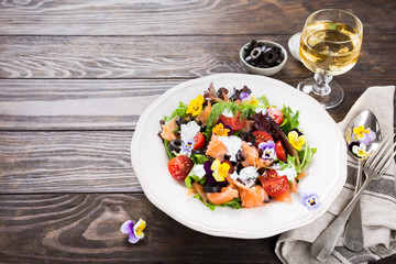 Fresh salad with smoked salmon, black olives, cherry tomatoes and edible flowers on wooden background.