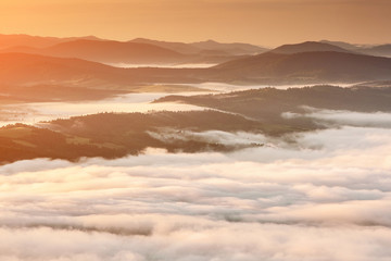 Fototapeta na wymiar Summer weather phenomenon. Seasonal landscape with morning fog in valley. Clouds drenched valley below the level of the mountains. Sunrise over creeping clouds.