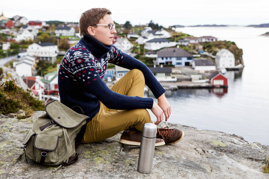 A young man sits on top of a hill on a background of ocean bay and a small Scandinavian town. Cloudy weather, autumn. Some men thermos of tea and a backpack.
