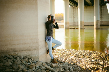 young woman in blue jeans and jacket, standing on the river bank with his back to the support of the bridge bent knee