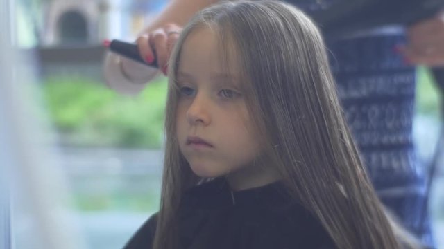 Hairdresser Combs and Dries Blonde Kid's Hairs Hairdresser Salon Stylist is Making a Haircut Hairstyle Little Girl is Sitting With Serious Face in Peignoir