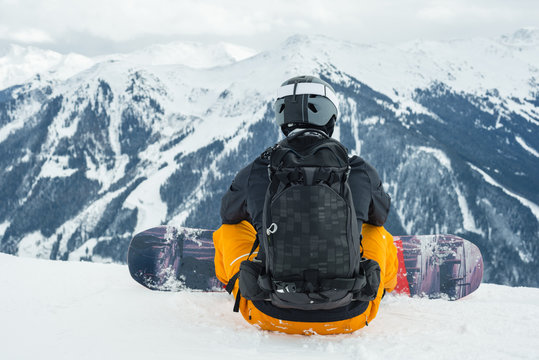 Rear view of man with snowboard sitting against snowcapped mountains