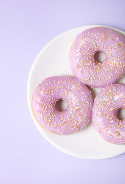 A plate of ring donuts with pastel pink frosting and sprinkles on a purple background