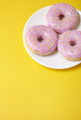 A plate of ring donuts with pastel pink frosting and sprinkles on a bright yellow background with blank space below