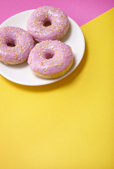 A plate of ring donuts with pastel frosting and sprinkles on a pink and yellow background with blank space below