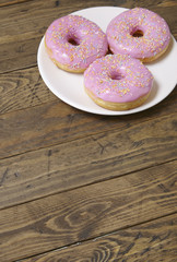A plate of ring donuts with pastel pink frosting and sprinkles on a rustic wooden table background with blank space below