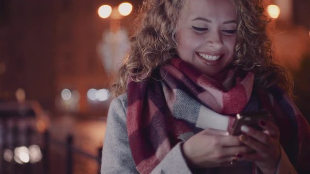 Attractive young woman in a warm grey coat and colorful scarf in the city street, surfing her phone and smiling. No people around. Female portrait. Rainy weather, city lights on the background.