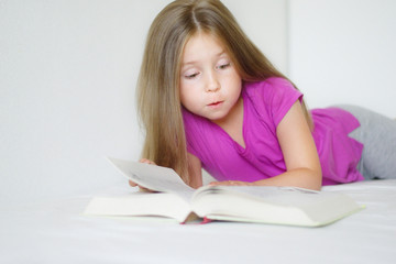 Adorable little girl lying on the bed and reading a book