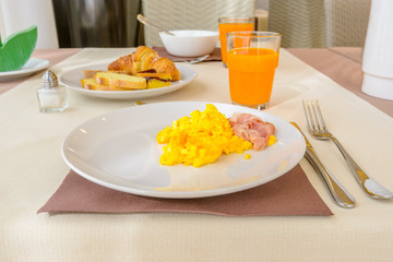 Breakfast including scrambled egg, becon, bread,  orange juice, and croissants.