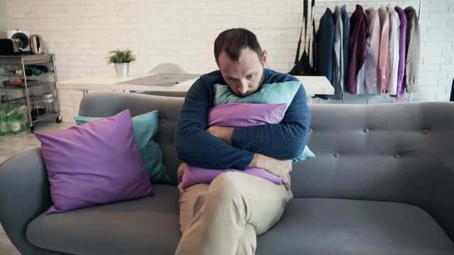 Sad, unhappy man hugging pillow sitting on sofa at home 
