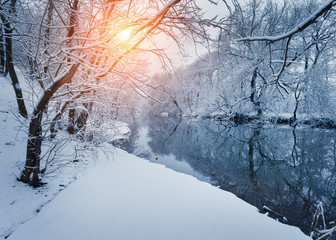 Winter forest on the river at sunset. Colorful landscape with snowy trees, frozen river with reflection in water. Seasonal. Winter trees, lake, sun and blue sky. Beautiful snowy winter in countryside