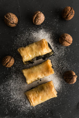 Three baklavas on stone black plate with suggar and walnuts around. Top view